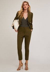 Oliwkowy, dopasowany garnitur z podwójnym zapięciem, slim-fit spodnie i błyszczący szary top, w zestawieniu z sandałami na wysokim obcasie z paskami.