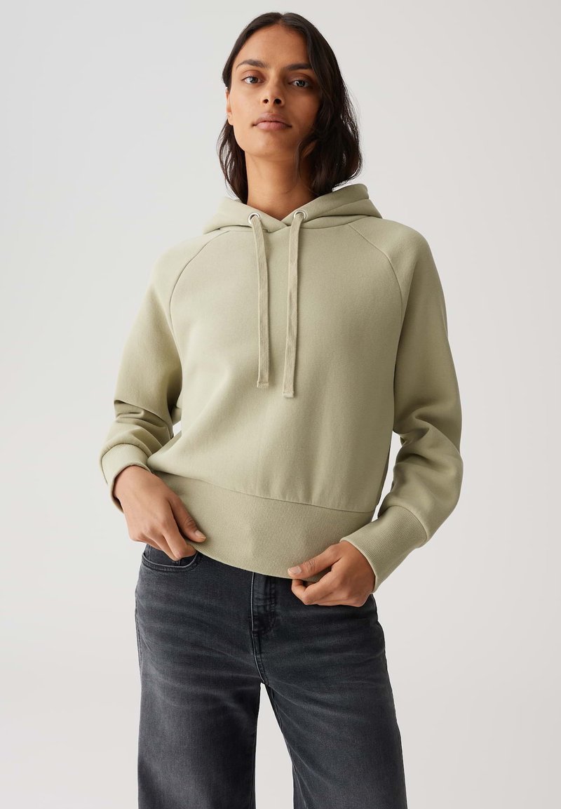 Donna che indossa una felpa con cappuccio verde chiaro e jeans neri, in piedi con le mani vicino alla vita su uno sfondo neutro.