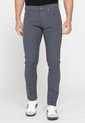 Uomo che indossa pantaloni aderenti grigio scuro e scarpe da ginnastica bianche, in piedi davanti a uno sfondo chiaro e uniforme.