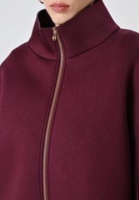 Burgunderfarbene Zip-Jacke mit hohem Kragen; hat eine glatte Textur und goldene Reißverschlussdetails entlang der vorderen Naht.