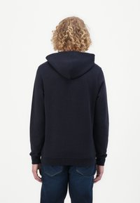 Felpa navy con cappuccio a stringa, tasca kangaroo, polsini e orlo a coste. Presenta una texture liscia con una vestibilità comoda. Jeans blu visibili sotto.