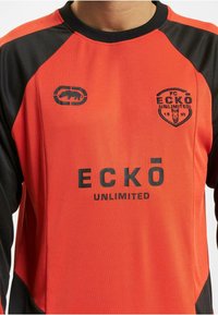 Rød sports skjorte med svarte detaljer, med en logo på venstre bryst og fet svart tekst som står "ECKÖ UNLIMITED." Lett stoff med glatt tekstur.