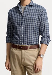 Modrá a bílá kostkovaná košile na knoflíky s vyhrnutými rukávy, hnědý kožený pásek a hodinky. Béžové kalhoty dokončují celý outfit.