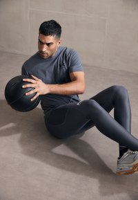 Sportos öltözet, amely egy szürke rövidujjú felsőt és sötétszürke leggingset tartalmaz, világosszürke és világosbarna cipővel párosítva, fitnesz környezetben.