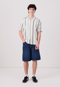 Chemise blanche à manches courtes et boutons, avec des rayures verticales noires, assortie à un short en denim bleu de longueur genou et des baskets noires.