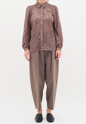Mujer con blusa marrón de encaje de manga larga, pantalones taupe holgados y zapatos negros sin cordones sobre un fondo blanco liso.