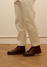 Bruine leren enkelboots met een gestructureerde afwerking, voorzien van vetersluiting en robuuste zolen. Gecombineerd met crèmekleurige maatpakken.