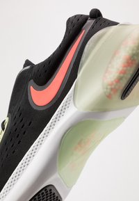 Zapatilla deportiva negra con un diseño texturizado, que presenta un logo de Nike en neón rosa y una entresuela verde translúcida con amortiguación visible.