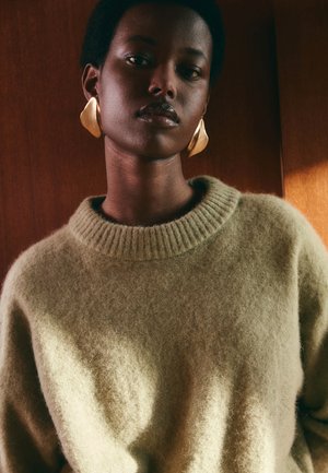 Femme aux cheveux courts portant de grandes boucles d'oreilles en or et un doux pull vert olive, se tenant devant un fond en bois avec un éclairage chaleureux.