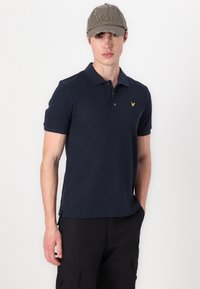 Jūras spēku polo krekls ar teksturētu audumu un dzeltenu logo kreisajā krūtī. Modelis valkā pelēku nolietotu cepuri ar tekstu "Won".