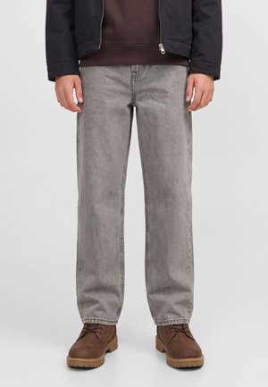 Modèle portant un jean gris ample, une veste foncée et des bottes marron à lacets, cadré de la taille aux pieds sur un fond uni.