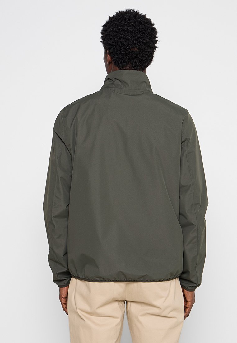 Giacca leggera verde con colletto alto e polsini elasticizzati. Texture liscia, vestibilità ampia e orlo con coulisse. Design minimalista, senza loghi visibili.