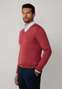 Czerwony sweter w serek wykonany z miękkiego materiału, dopasowany krój, z długimi rękawami i ściągaczami, nałożony na jasnoniebieską koszulę z kołnierzykiem.