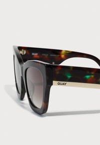 Gafas de sol de carey con montura cuadrada, lentes oscuros degradados, acentos dorados en la varilla y el logo "QUAY" en el lateral.