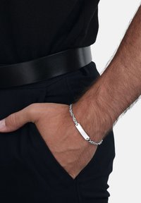 Męska dłoń w kieszeni ciemnych spodni, nosząca srebrną bransoletkę i czarny pasek z czarną koszulą na tle jednolitym.