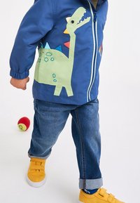 Giacca blu per bambini con un'applicazione di un dinosauro verde sul lato, dotata di spuntoni multicolori. Abbinata a jeans di denim e scarpe gialle.