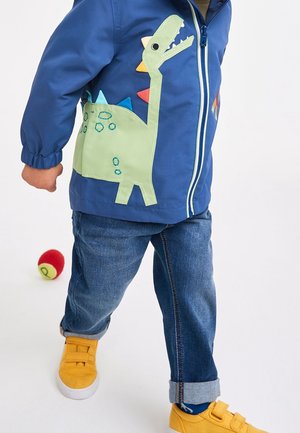 Blue children's jacket featuring a green dinosaur appliqué on the side, with multicolored spikes. Paired with denim jeans and yellow shoes.