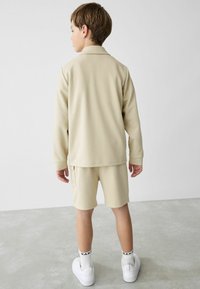 Beige, teksturoitu takki ja shortsiyhdistelmä. Takissa on kaulus ja pitkät hihat; shortsit ovat polven yläpuolella. Valkoiset lenkkarit ja kuvioidut sukat.