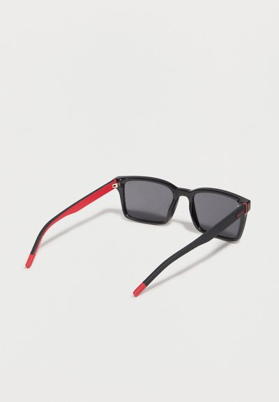 Lunettes de soleil noires carrées avec des verres foncés, des branches intérieures rouges et des branches extérieures texturées noires reposant sur une surface blanche.