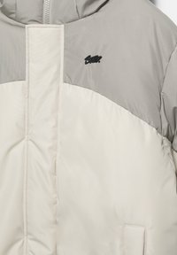 Letvægts, tofarvet dunjakke med en cremefarvet nederste del og grå øverste del. Har et broderet mønster og et lille sort logo.