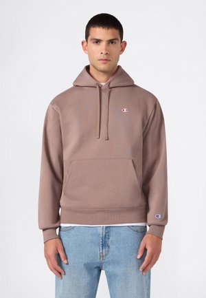 Mann trägt braunen Champion-Hoodie mit Fronttasche und hellblauen Jeans und steht vor einem einfarbigen weißen Hintergrund.