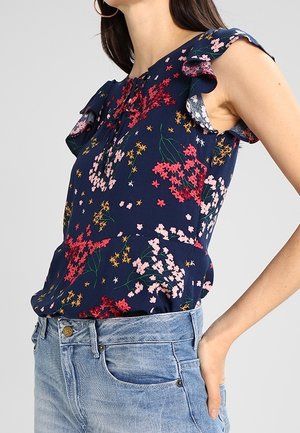 Laivastonsininen hihaton pusero, jossa on punaisen, vaaleanpunaisen ja keltaisen kukkakuvio, röyhelöidyt olkapäät, yhdistetty vaaleansinisin farkkuihin.