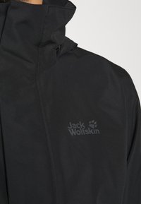 Veste imperméable noire avec col montant et logo Jack Wolfskin sur la poitrine gauche, portée par une personne.