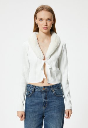Weißes geripptes kurzes Cardigan mit einem flauschigen weißen Kragen, der vorne gebunden ist. Kombiniert mit hochgeschnittenen blauen Jeans.