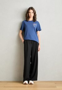 Blue cotton t-shirt with short sleeves and white graphic print; paired with loose-fitting black trousers and white sneakers.