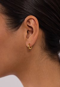 Goldene Creole, die am Ohr eines Models präsentiert wird, mit einem glatten, glänzenden Finish, kreisförmiger Form und kleinem, kompakten Design.