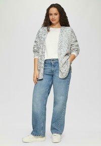 Lichtgrijze cardigan met gestructureerde spikkels, gedragen over een wit topje. Gecombineerd met blauwe wijde jeans en witte sneakers, handen in de zakken.
