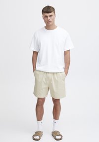 Tricou alb cu mânecă scurtă, pantaloni şort bej cu bandă elastică la talie, şosete albe şi sandale de tip slide maro. Texturi simple şi moi.