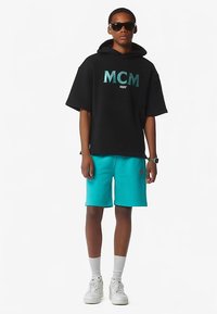 Zwarte oversized hooded sweatshirt met turkooizen "MCM" logo, gecombineerd met turkooizen shorts en witte sneakers. Casual atletische stijl.