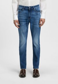 Blue denim jeans with a straight fit, five-pocket design, subtle fade, zip fly, and button closure, worn with brown leather shoes.