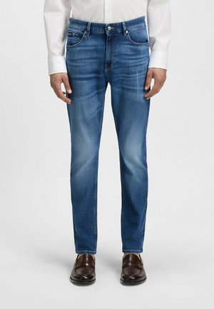 Blue denim jeans with a straight fit, five-pocket design, subtle fade, zip fly, and button closure, worn with brown leather shoes.