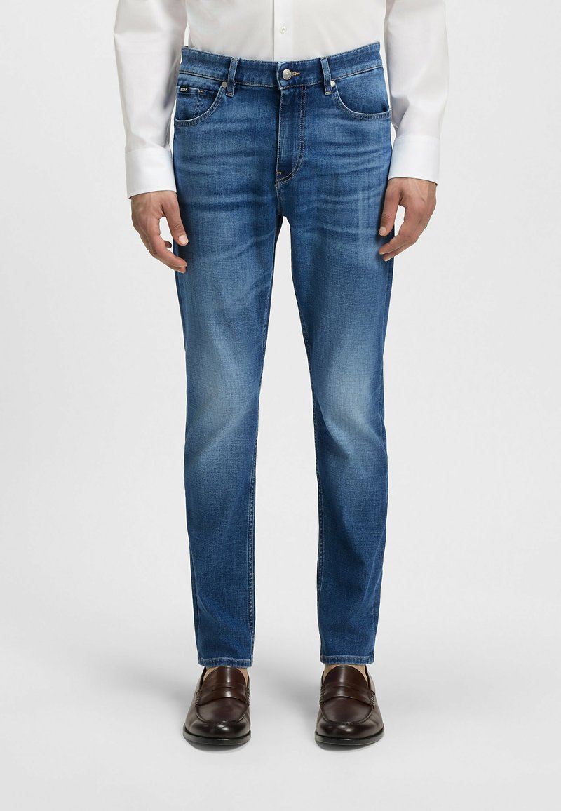 Blue denim jeans with a straight fit, five-pocket design, subtle fade, zip fly, and button closure, worn with brown leather shoes.