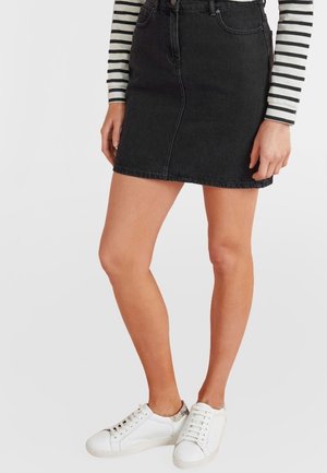Femme portant une mini-jupe en denim noire, un haut à manches longues rayé et des baskets blanches avec un détail à motifs sur le talon, debout devant un fond uni.
