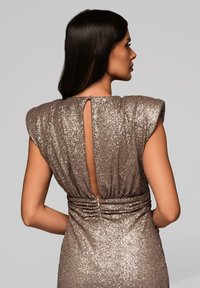 Vrouw met stijl donker haar draagt een mouwloze, glinsterende taupe jurk met een verticale uitsnijding aan de achterkant en gerimpelde taille, staande in profiel.