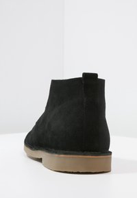 Bottine noire en daim avec une texture lisse, semelle en caoutchouc beige contrastante et un design simple et épuré. Pas de matériel ou de motifs visibles.