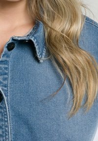 Denim shirt with a light blue color, featuring a collared neck, black buttons, and visible stitching along the edges. Soft texture.