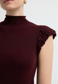 Top de punto burdeos con cuello alto y mangas cortas, con acentos de volantes. Textura suave con un diseño ajustado. Pendientes de oro visibles.