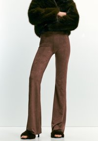 Persona che indossa pantaloni scampanati in suede marrone, sandali con tacco pelosi neri e una giacca in pelliccia sintetica verde scuro, con le braccia incrociate.