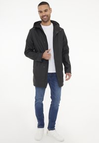 Chaqueta negra impermeable con capucha, que cuenta con un cierre de presión en el frente y bolsillos laterales, combinada con unos jeans azules y zapatillas blancas.