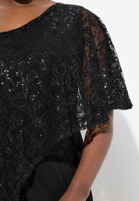 Top de encaje negro con un diseño fluido, que presenta patrones intrincados y lentejuelas. Mangas cortas y anchas y una capa negra suave por debajo.