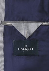 Unutrašnjost tamnoplave jakne s trakastim rubom od tkanine i pravokutnom crnom oznakom na kojoj piše "HACKETT LONDON" sašivenom iznutra.