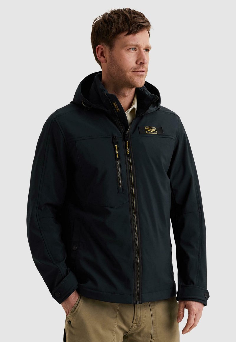 Mann trägt eine schwarze Kapuzenjacke mit durchgehendem Reißverschluss und Brusttasche, kombiniert mit beigen Hosen, und schaut nach rechts.