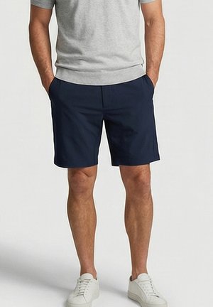 Mann, der marineblaue Shorts, graues T-Shirt und weiße Sneaker trägt, steht mit den Händen in den Taschen vor einem einfarbigen Hintergrund.