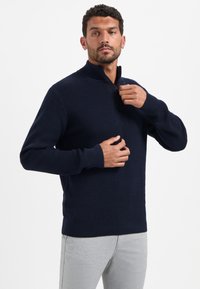 Man in een marineblauwe wajif gebreide half-rits trui en lichtgrijze broek, die de rits van de kraag van de trui aanpast.