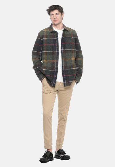 Uomo in piedi con le mani in tasca, indossa un giubbotto a quadri verde e nero, una camicia bianca, pantaloni beige e scarpe eleganti nere.
