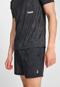 Czarny strój sportowy składający się z pasiaka z krótkim rękawem i dopasowanych szortów, obie części o gładkiej fakturze i kontrastowym białym logo.